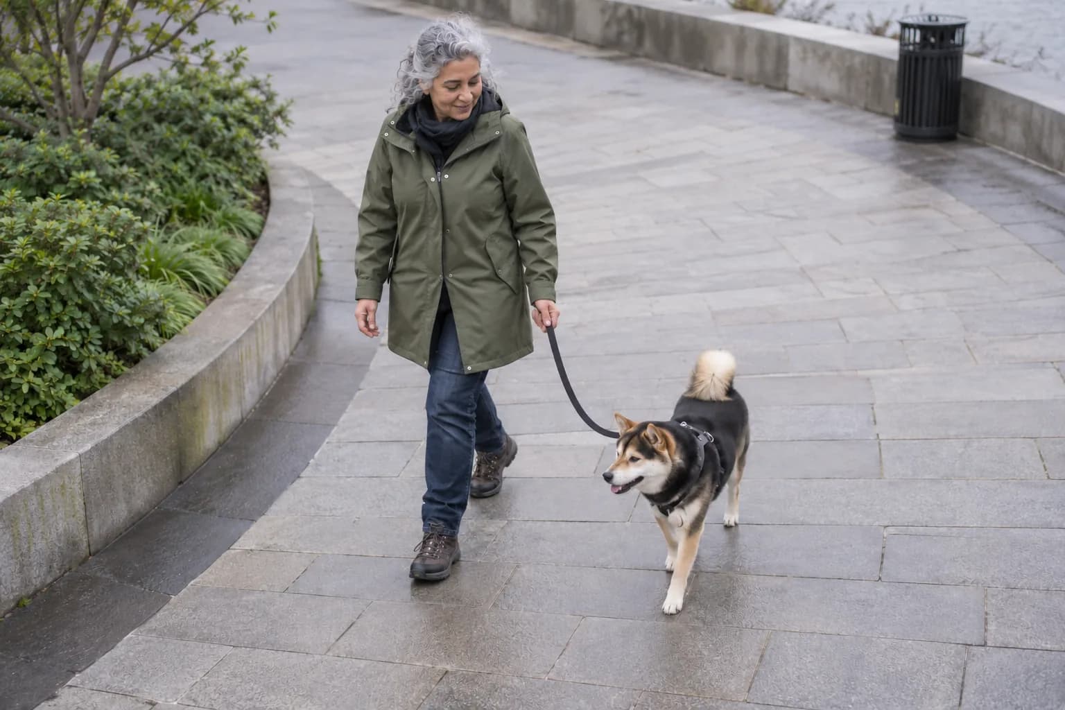 A woman and her leashed Shiba Inu follow a gentle curve across empty paving away from a distant closed bin